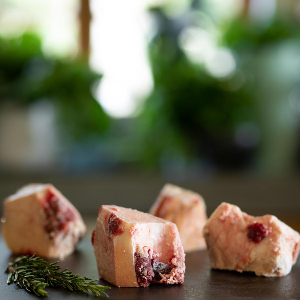 Several raw frozen meaty beef bones on a dark surface, with a sprig of rosemary to the side.